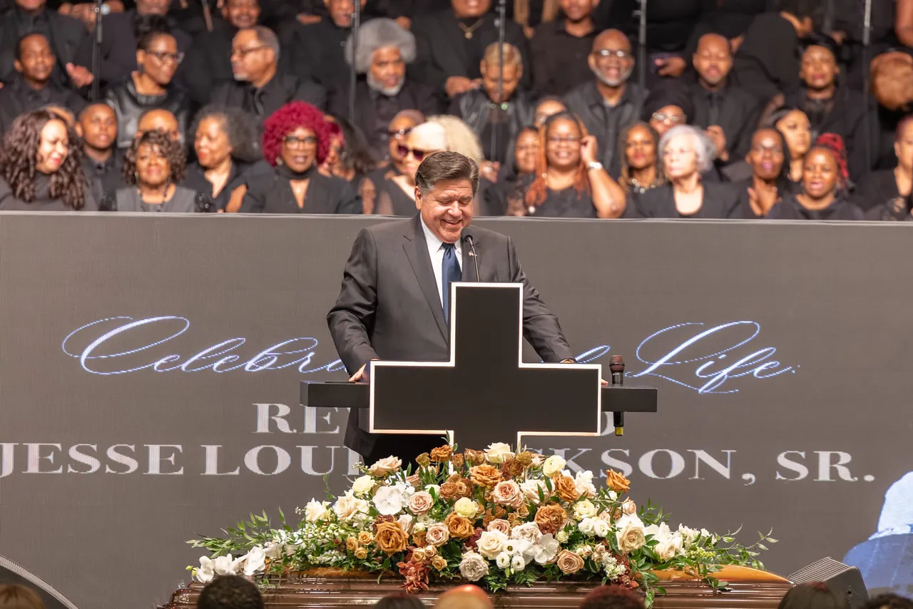 A speaker at the lectern behind a spray of flowers, the choir in black behind him