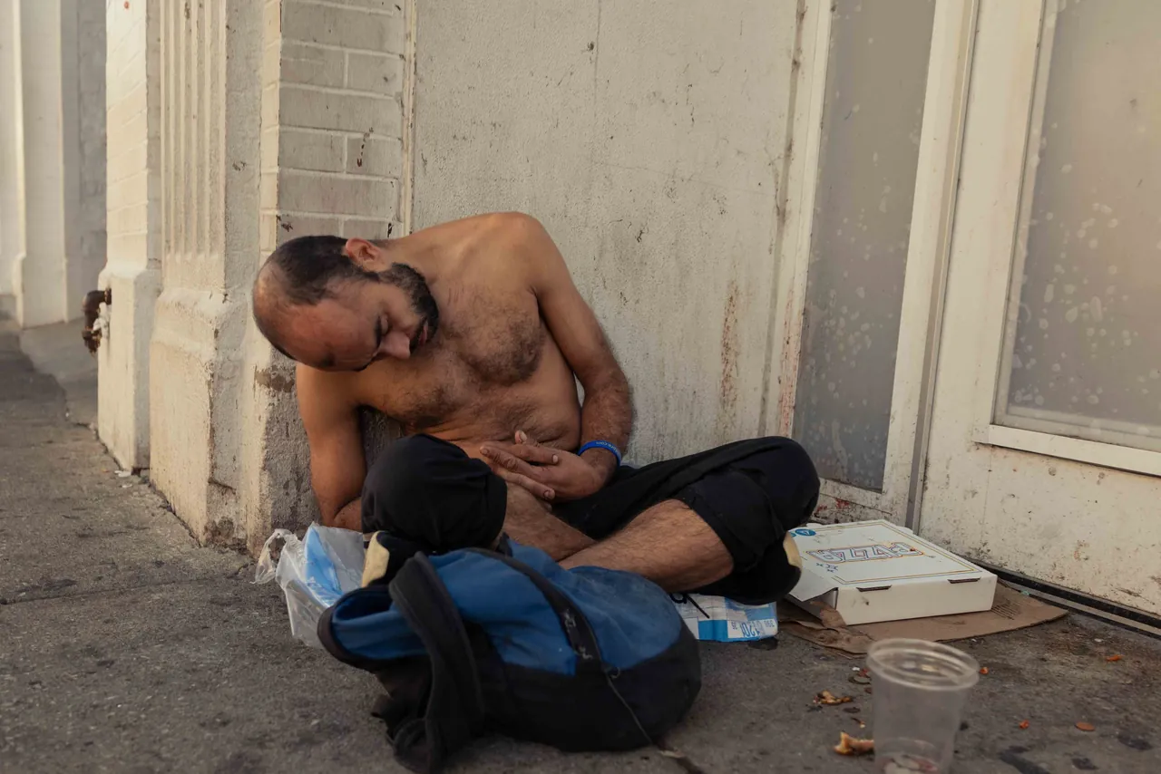 A shirtless man sits against a wall on the sidewalk
