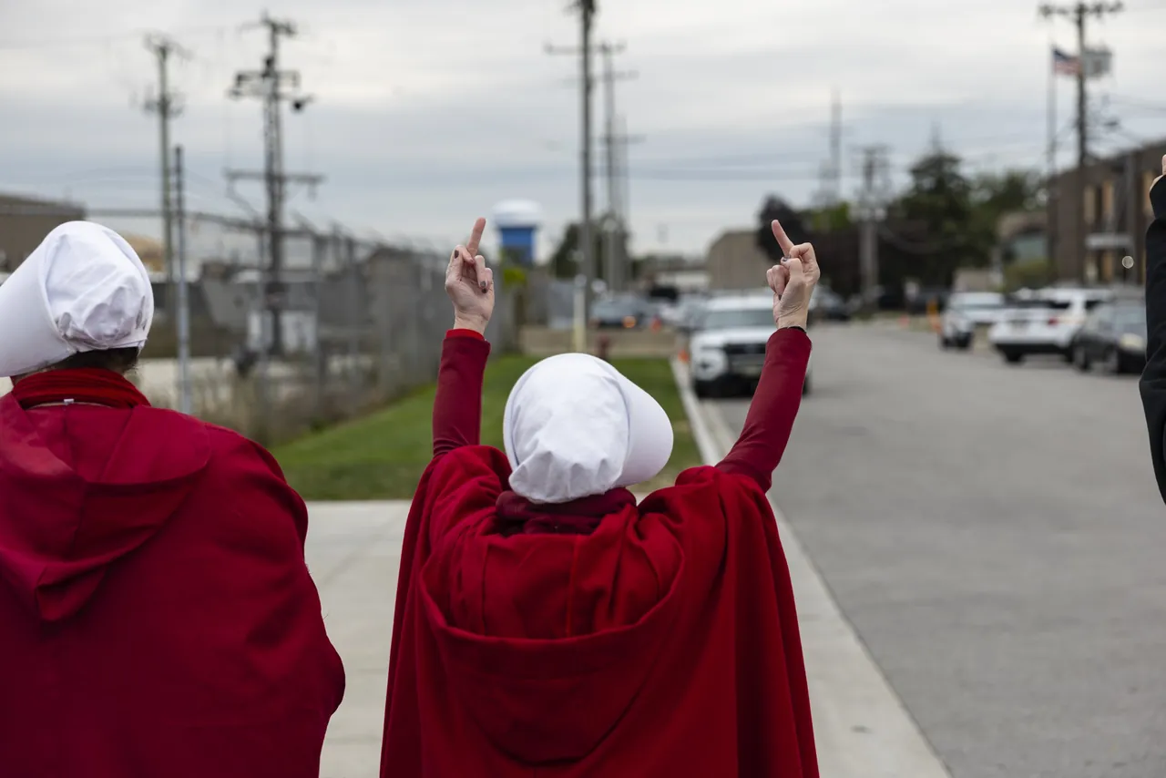 Two demonstrators in red cloaks and white bonnets raise their middle fingers toward a fenced lot