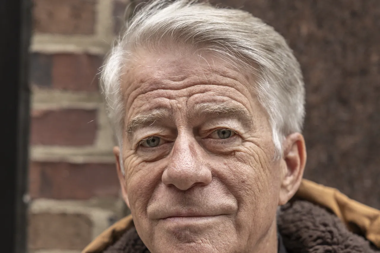 Close portrait of an older man with silver hair in a tan coat against brick